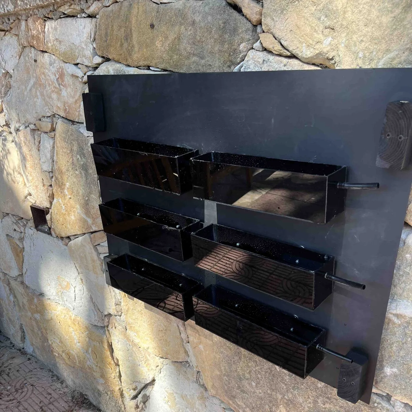 Six empty black planters hanging in three rows on the back panel, on a stone wall.